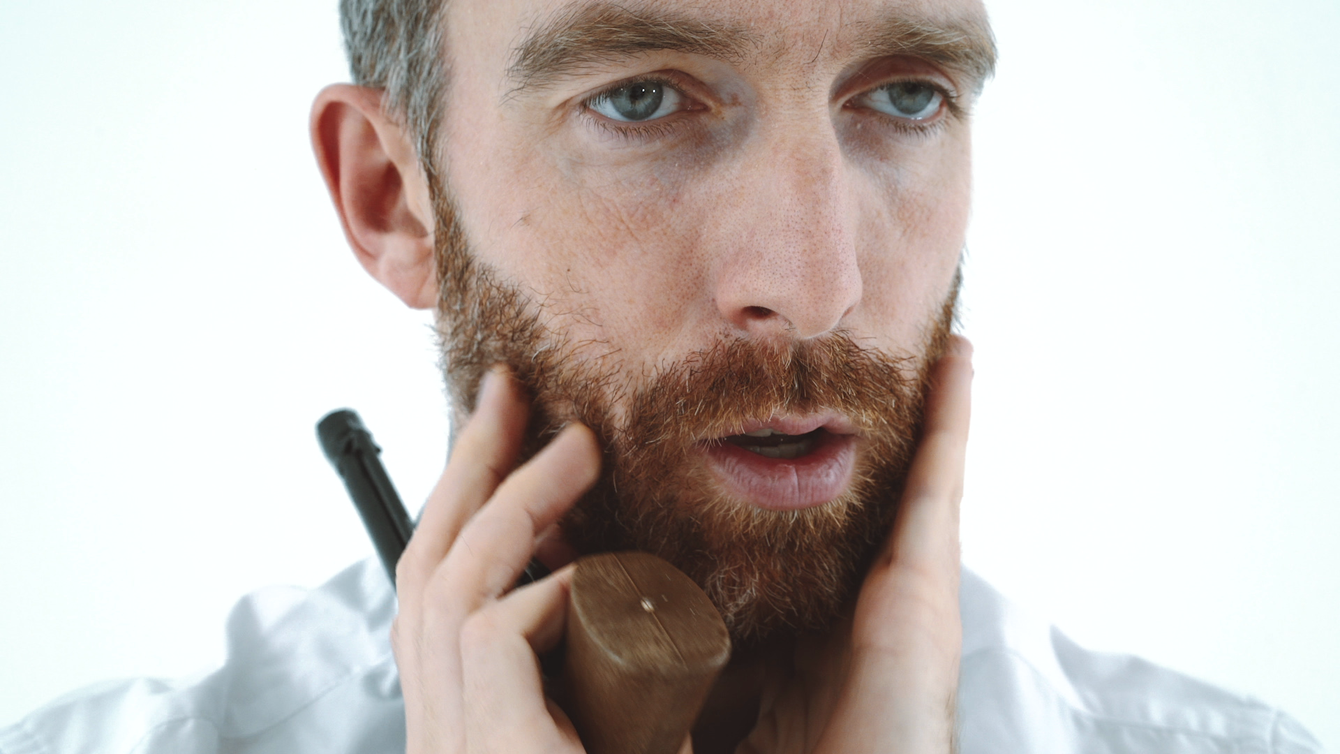 A man with a thoughtful expression holds his face and a wooden pipe. He has a beard, wears a white shirt, and seems deep in contemplation.