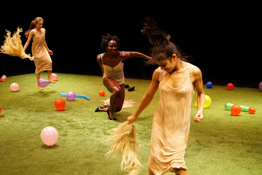 Three women in beige dresses dance joyfully on a grass stage, surrounded by colourful balloons. The scene conveys energy and playfulness.