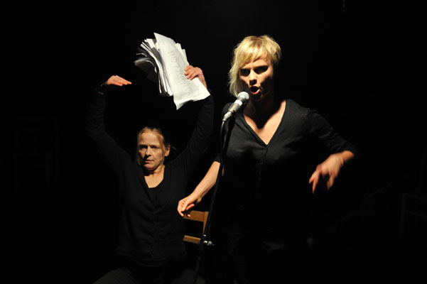 Two performers dressed in black on a dimly lit stage. The one in front passionately speaks into a microphone, while the other raises papers dramatically.