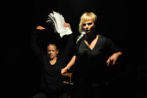 Two performers in dramatic lighting; one passionately sings into a microphone, the other energetically holds up papers, both in black attire on a dark stage.