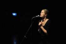 A woman in a dimly lit setting stands at a microphone, looking upwards with a hopeful expression, hands clasped. The background is dark, creating an emotional ambiance.