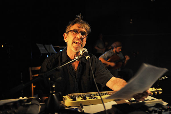 A man in a dark shirt passionately sings into a microphone, holding sheet music. A guitarist plays in the dimly lit background, creating an intimate atmosphere.