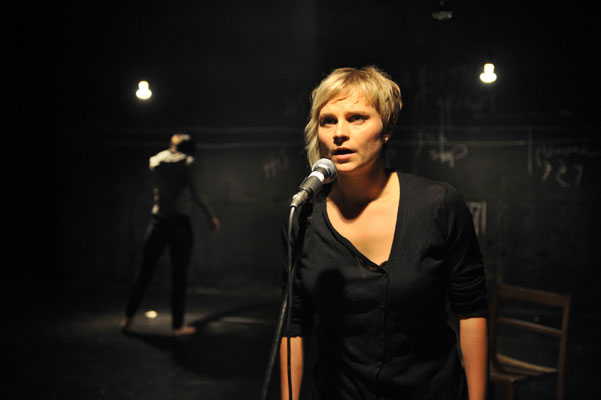 A woman stands at a microphone on a dimly lit stage, wearing a black shirt, appearing focused. In the background, another person is barely visible, turned away. The mood is intense and dramatic.