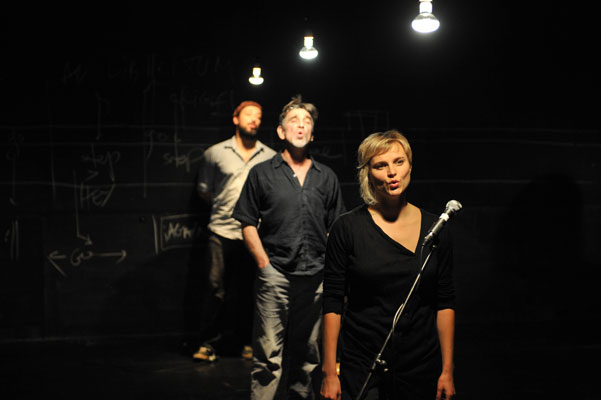 A woman sings into a microphone on a dimly lit stage, with two men standing behind her. Chalk diagrams on a blackboard create an artistic backdrop.
