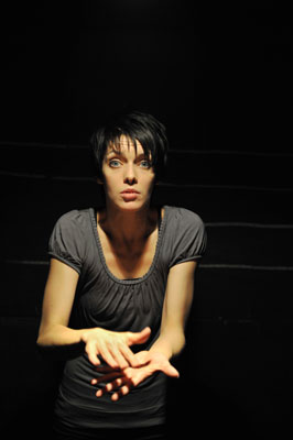 A woman with short hair and a serious expression stands in dim lighting, reaching her hands forward. The scene conveys a tense and dramatic mood.