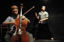 Man playing cello on stage in soft lighting, focused, while another man stands in the background with a poised, dramatic pose. Artistic performance scene.