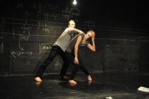 Two barefoot dancers perform in a dimly lit space with chalk diagrams on a blackboard. One supports the other in a dramatic, leaning pose.