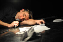 Man lying on a dark stage, holding a microphone, surrounded by scattered papers. His facial expression is contemplative and introspective.
