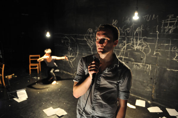 A man stands in dim light holding a microphone, with a serious expression. Behind him, another person moves dynamically near scattered papers and chalk drawings on a dark stage.
