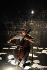 A man plays a cello on a dimly lit stage, surrounded by scattered papers. Chalk drawings cover the black wall behind, creating an artistic, moody atmosphere.