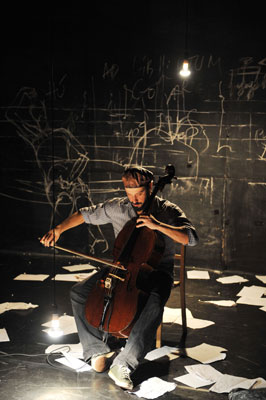 A man plays a cello on a dimly lit stage, surrounded by scattered papers. Chalk drawings cover the black wall behind, creating an artistic, moody atmosphere.