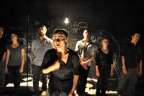A group of people stand on a dimly lit stage with chalkboard walls. One person in the foreground gestures passionately, creating an intense, dramatic atmosphere.