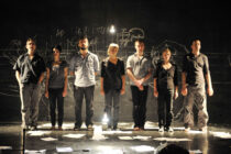 Seven people stand in a line on a dimly lit stage, with papers scattered on the floor and chalk drawings on the black backdrop, creating a sombre mood.
