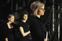 A woman with short blond hair, dressed in black, stands in profile holding a notebook. Two blurred figures in black are in the background. The scene conveys focus and anticipation.