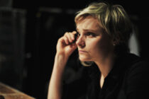 A woman with short blonde hair sits in a dimly lit room, looking thoughtfully to the side. She rests her hand near her face, conveying a contemplative mood.