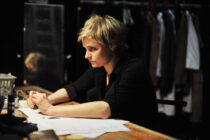 A focused woman with short blonde hair sits at a cluttered wooden table, reviewing papers. The dimly lit room and hanging clothes suggest a backstage setting.