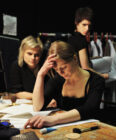 Three people backstage: a woman in front holds her head, focused on papers; two others watch, one looking directly at the viewer. Serious, concentrated mood.