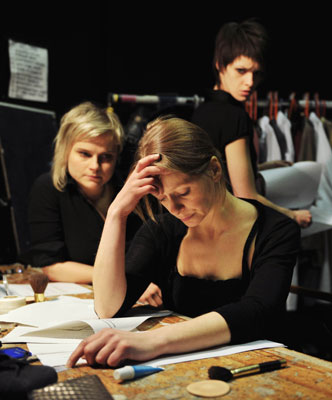 Three people backstage: a woman in front holds her head, focused on papers; two others watch, one looking directly at the viewer. Serious, concentrated mood.