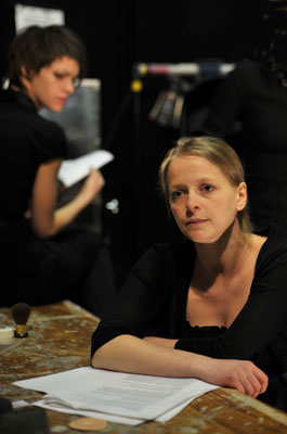A woman with blonde hair in a black top leans on a table with papers, looking pensive. Another person is blurred in the background, creating a contemplative mood.