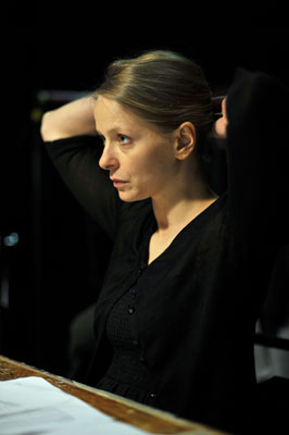 A woman in a black outfit is sitting at a table in a dark room, holding her hair up. She looks focused and thoughtful, with papers in front of her.