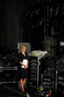 A woman in a black dress stands backstage, holding papers, surrounded by technical equipment and cables. The scene conveys anticipation and readiness.