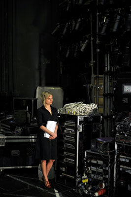 A woman in a black dress stands backstage, holding papers, surrounded by technical equipment and cables. The scene conveys anticipation and readiness.
