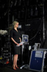 A woman in a black dress stands backstage, holding papers, surrounded by technical equipment and cables, appearing focused and composed.