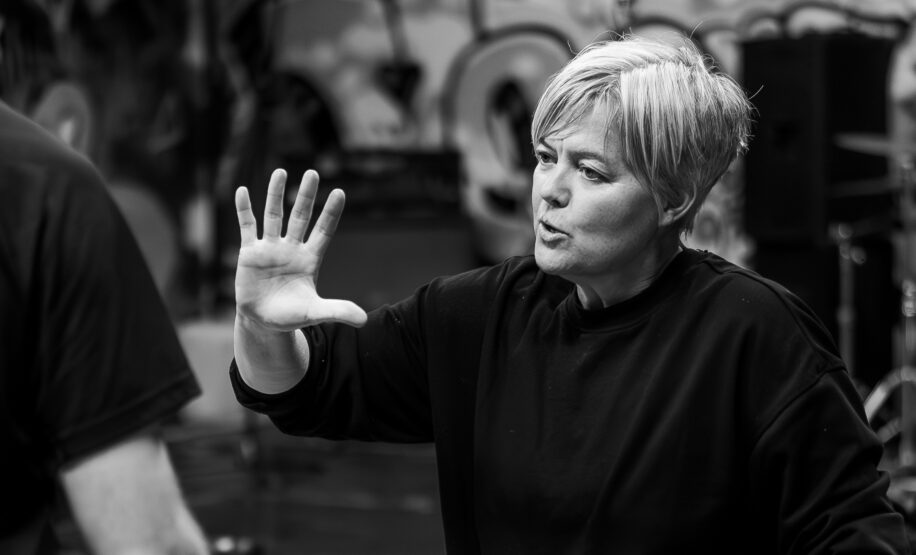A person in a black sweatshirt gesturing during a rehearsal in a vibrant, graffiti-filled space, with instruments visible in the background.