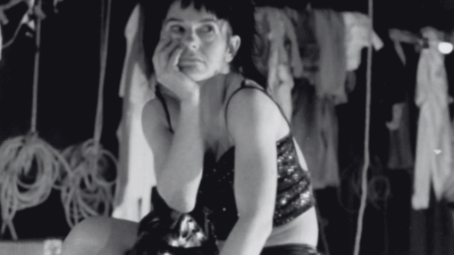 A performer sits pensively in a sequined outfit, surrounded by theatrical props in a dimly lit backstage setting.