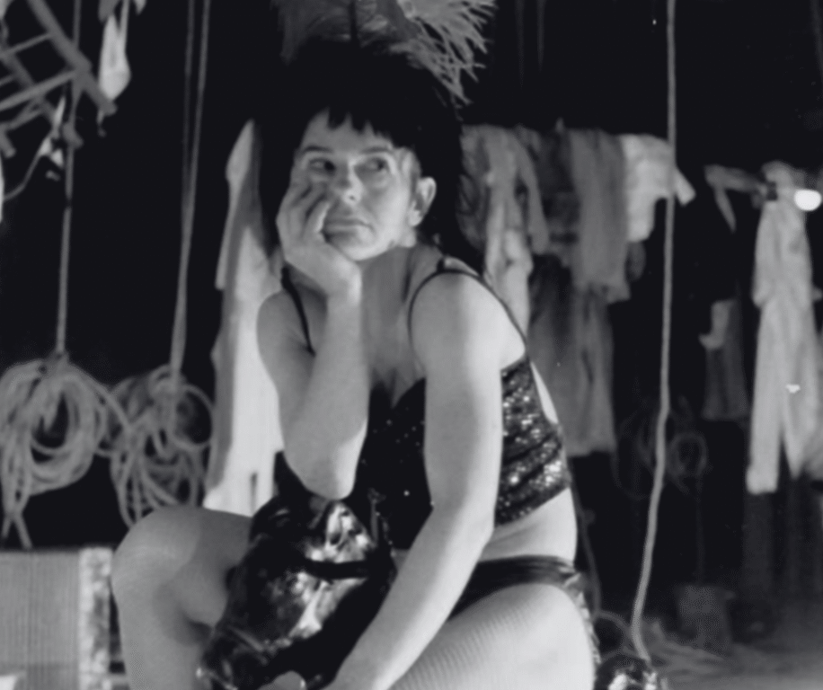 A performer sits pensively in a sequined outfit, surrounded by theatrical props in a dimly lit backstage setting.