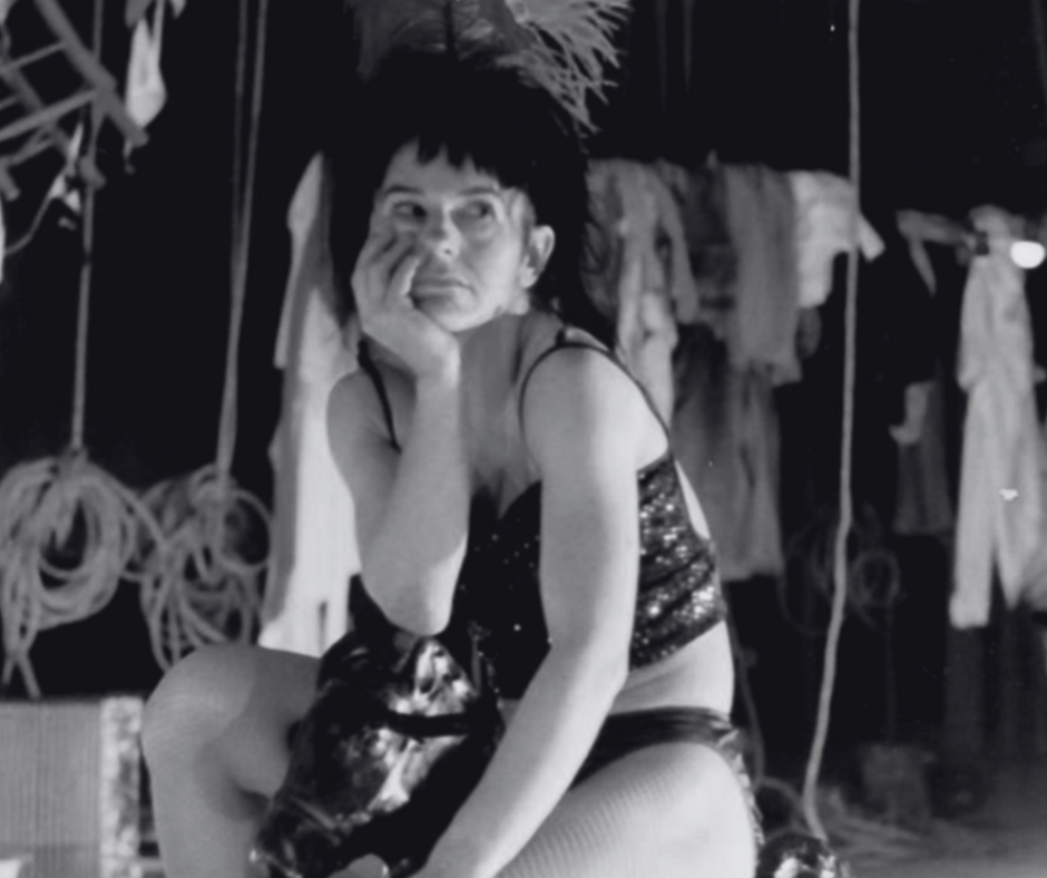 A performer sits pensively in a sequined outfit, surrounded by theatrical props in a dimly lit backstage setting.