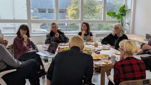 A group of nine people engaged in discussion around a table with laptops, papers, and snacks, illuminated by natural light from large windows. Casual and collaborative atmosphere.