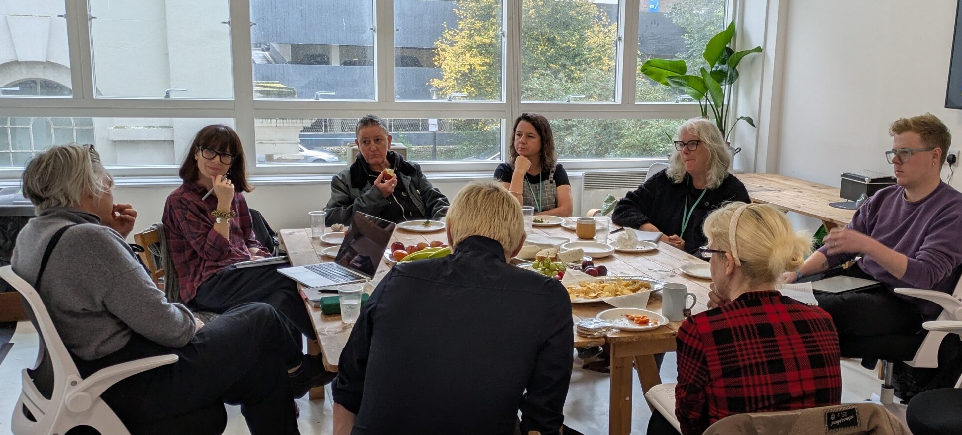 A group of nine people engaged in discussion around a table with laptops, papers, and snacks, illuminated by natural light from large windows. Casual and collaborative atmosphere.