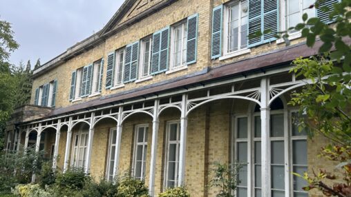 Historic building with light brick walls and turquoise shutters. White arches support a porch, surrounded by lush greenery under a cloudy sky.