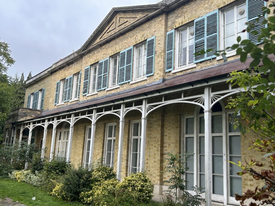 Historic building with light brick walls and turquoise shutters. White arches support a porch, surrounded by lush greenery under a cloudy sky.