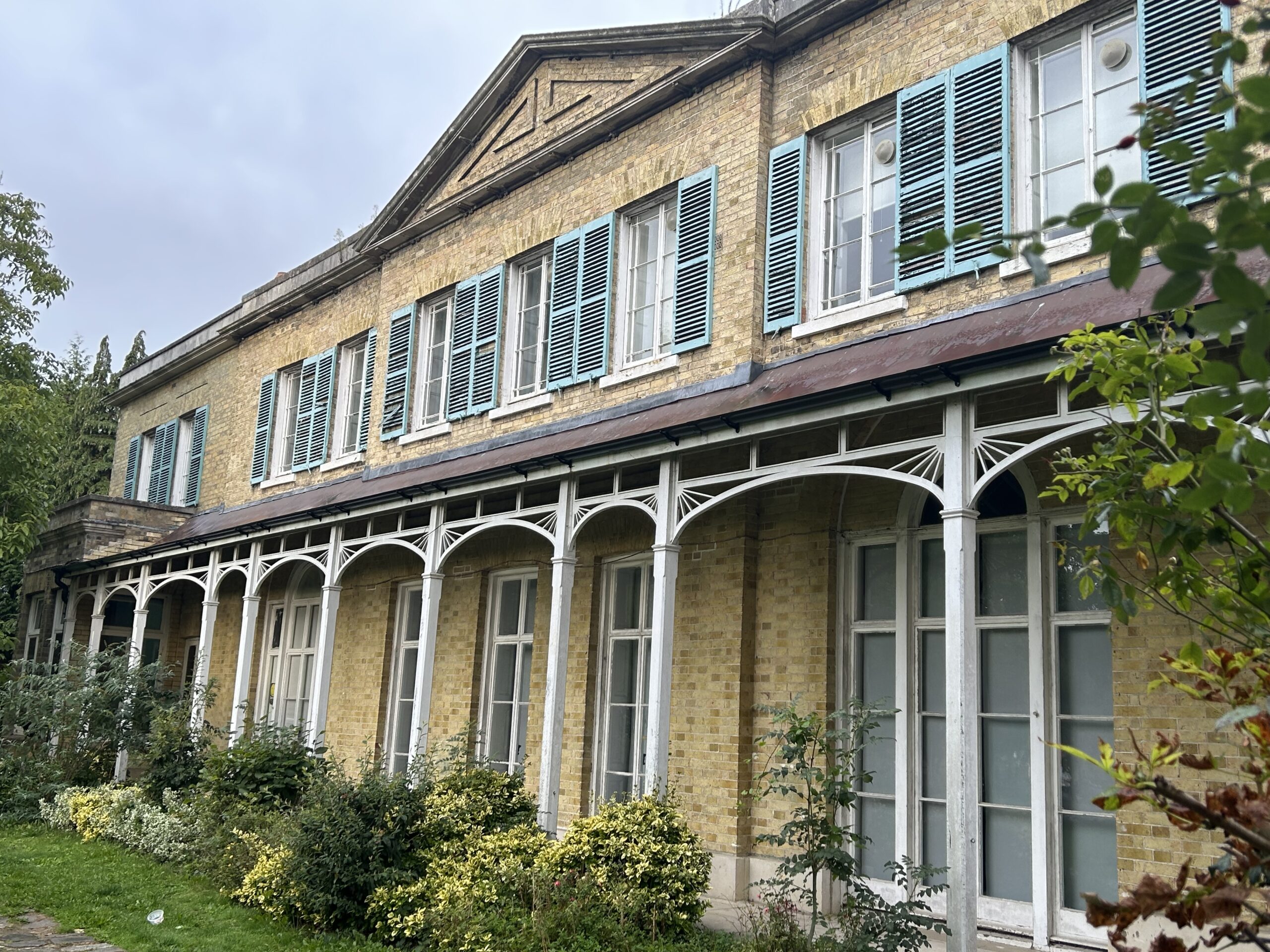 Historic building with light brick walls and turquoise shutters. White arches support a porch, surrounded by lush greenery under a cloudy sky.