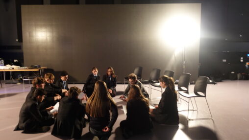 A group of people in casual attire sit in a circle on a studio floor under bright lighting. Empty chairs surround them, suggesting a collaborative setting.