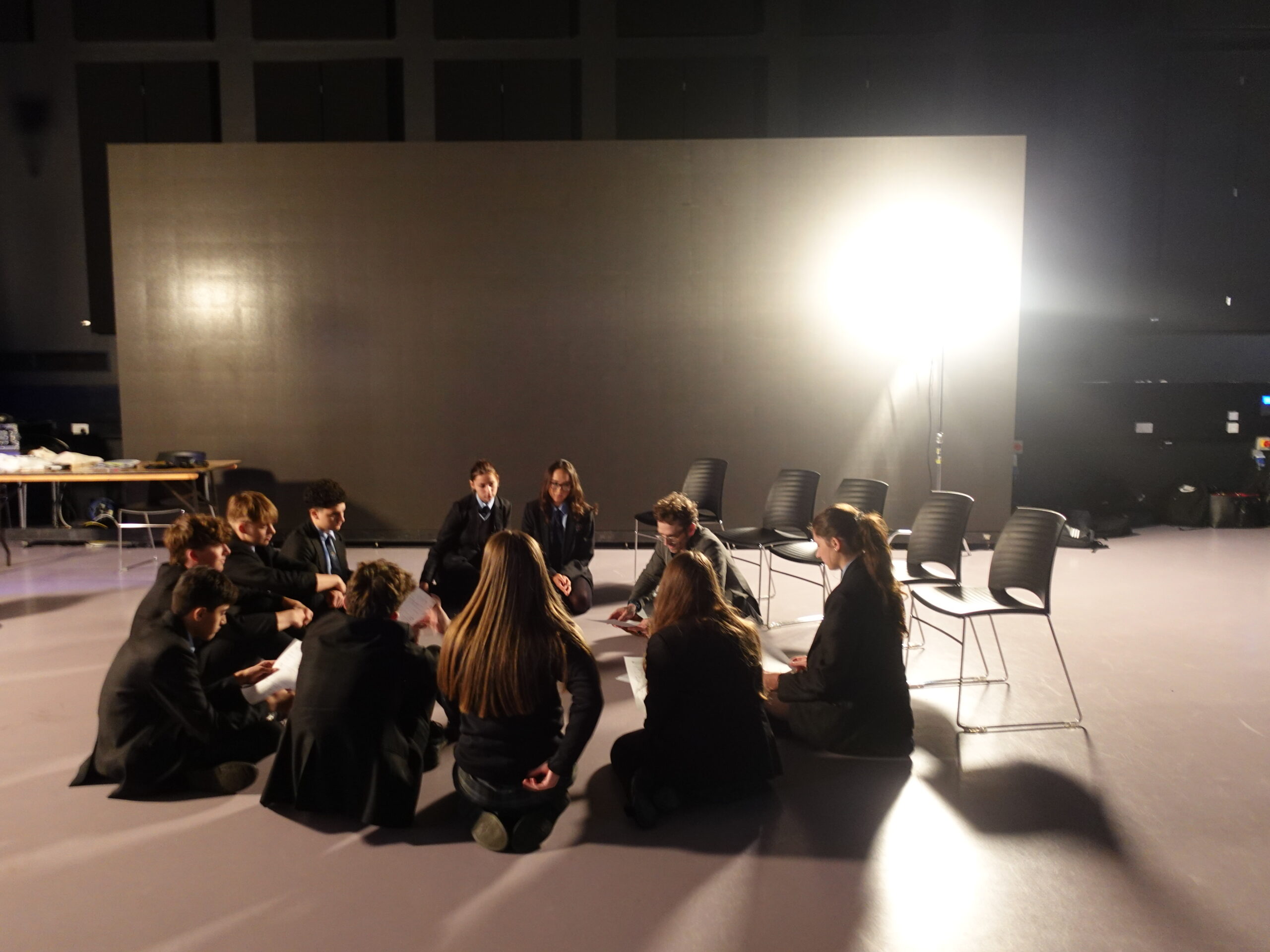 A group of people in casual attire sit in a circle on a studio floor under bright lighting. Empty chairs surround them, suggesting a collaborative setting.