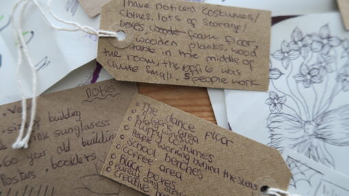 Close-up of several brown paper tags with handwritten notes in black ink. Tags are scattered on a table, with nature-themed sketches partially visible underneath.
