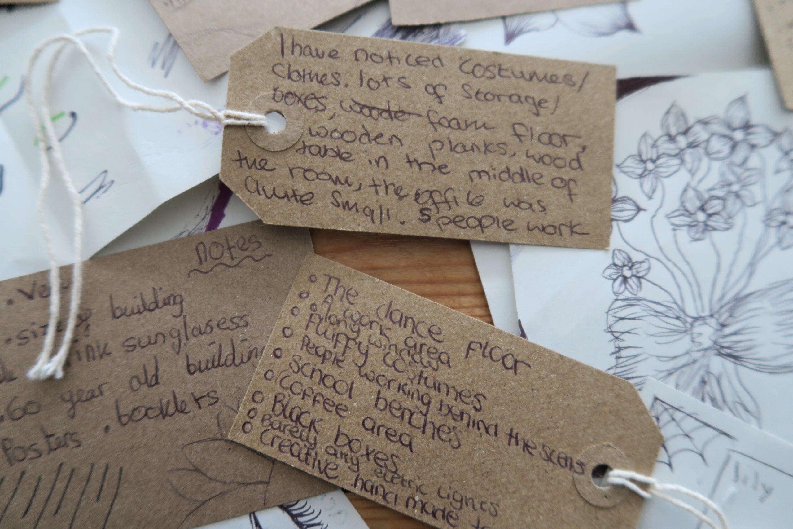 Close-up of several brown paper tags with handwritten notes in black ink. Tags are scattered on a table, with nature-themed sketches partially visible underneath.