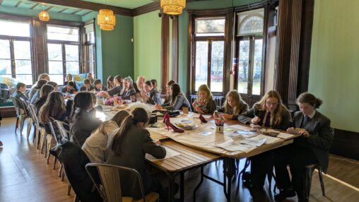 A group of students in uniforms sit around a large wooden table in a warmly lit room with large windows. They are focused on crafting projects.