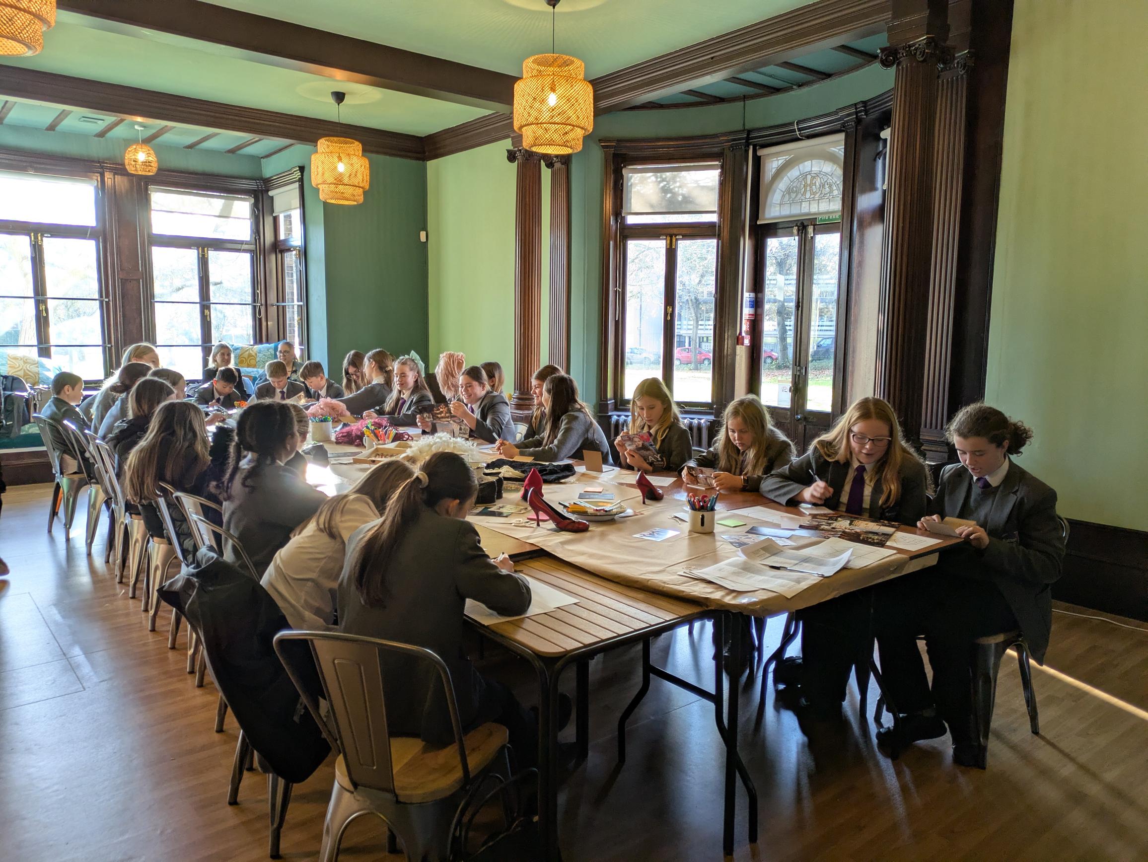 A group of students in uniforms sit around a large wooden table in a warmly lit room with large windows. They are focused on crafting projects.