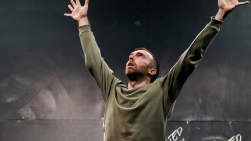 A man in a green sweater stands with arms raised against a dark background. Chalk scribbles are visible, conveying determination and intensity.