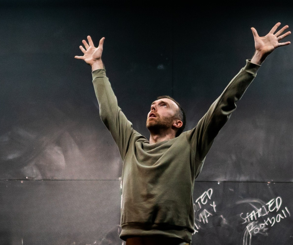 A man in a green sweater stands with arms raised against a dark background. Chalk scribbles are visible, conveying determination and intensity.