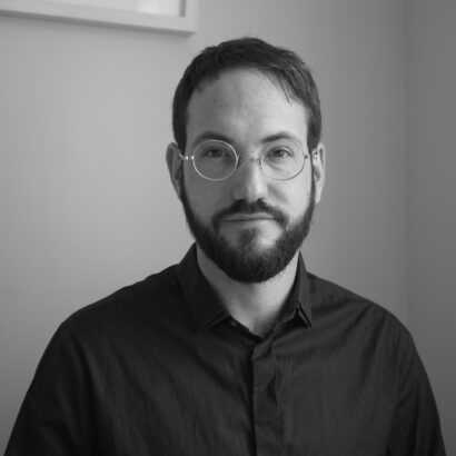 A man with short dark hair and a beard, wearing glasses and a navy blue shirt, looks at the camera in a neutral expression. The setting is a plain, light-coloured room.