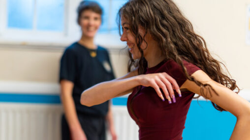 A woman in a burgundy top and red checked trousers dances energetically in a studio, while another person in black watches with a smile, creating a joyful atmosphere.