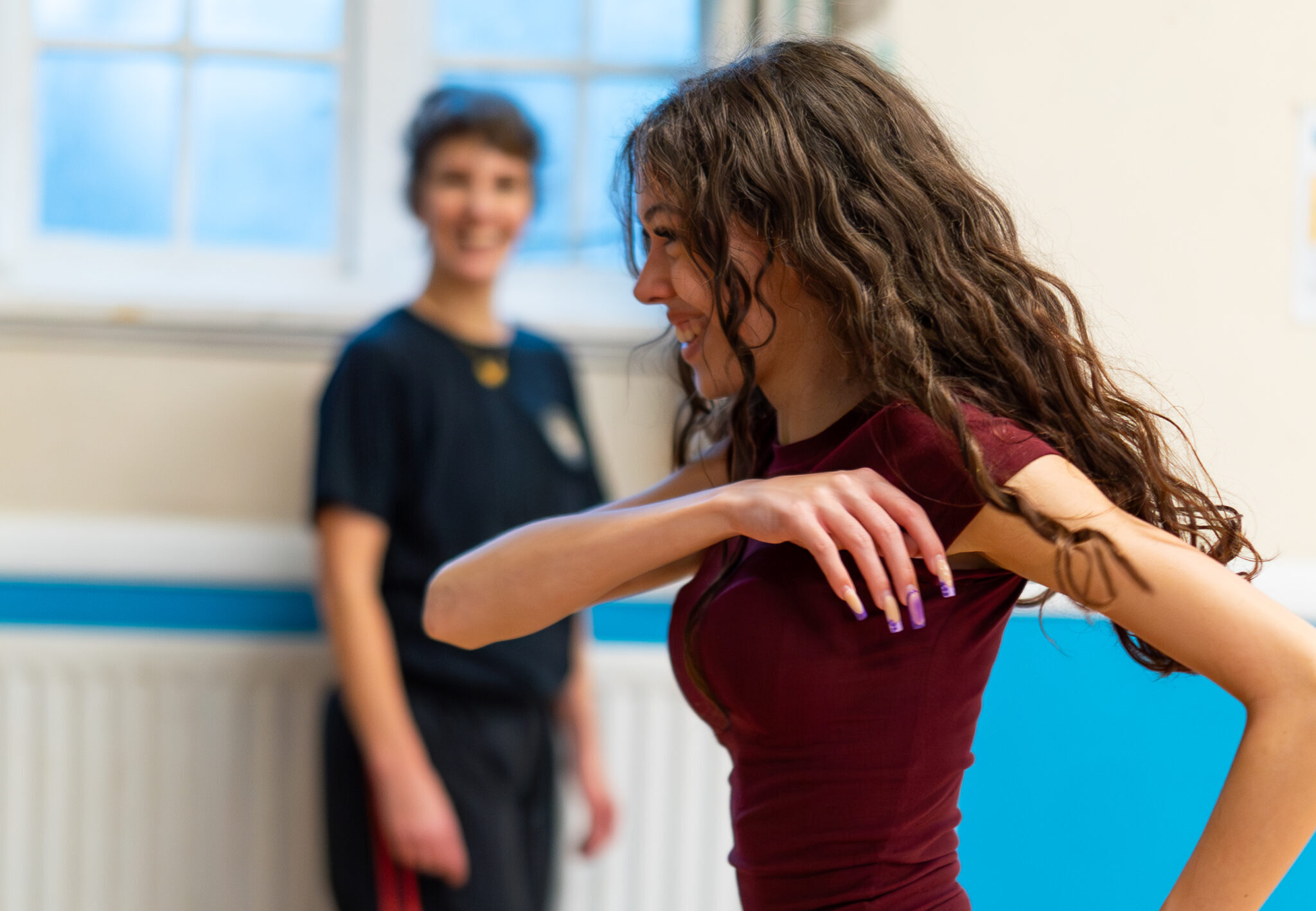 A woman in a burgundy top and red checked trousers dances energetically in a studio, while another person in black watches with a smile, creating a joyful atmosphere.