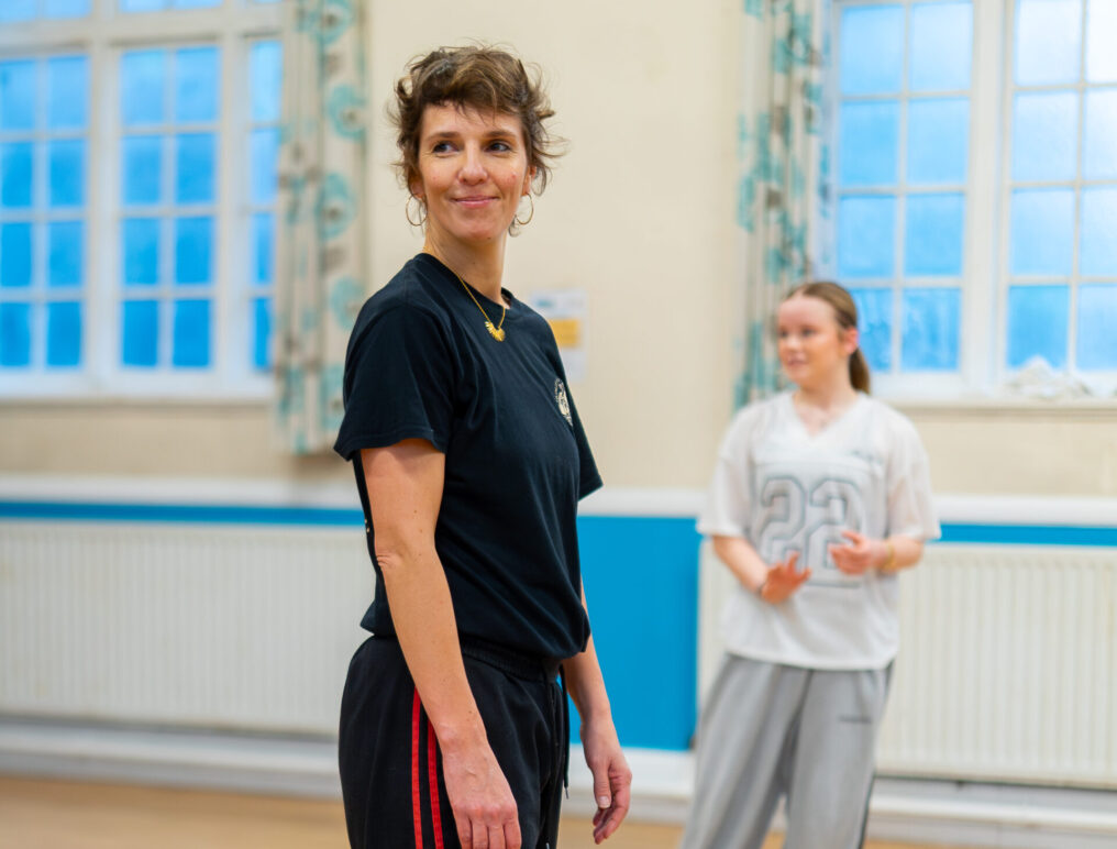 Woman in a dance studio wearing casual black athletic wear with red stripes, looking content. Another person in the background appears in motion.