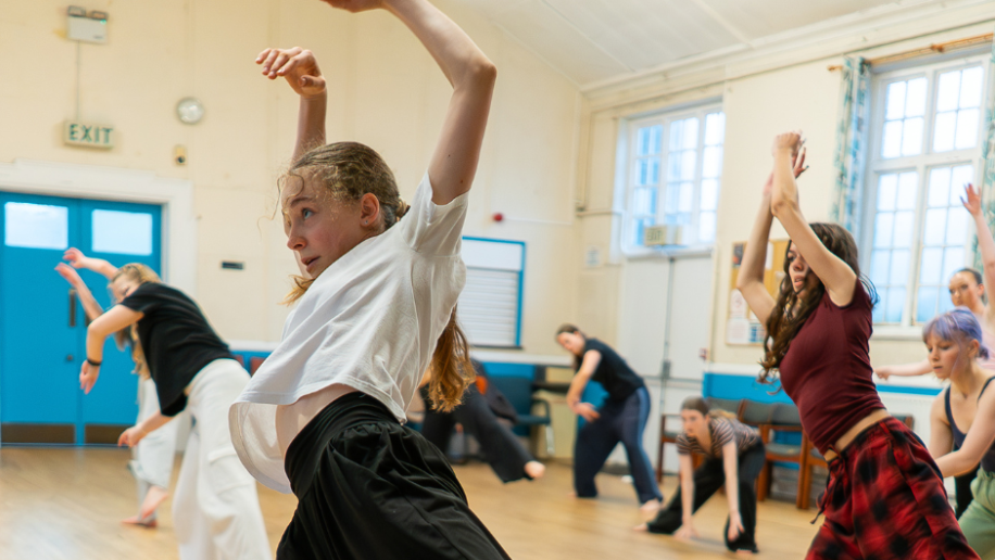 A group of people energetically dancing in a bright studio. The focus is on a person in a white shirt, expressing dynamic movement and concentration.