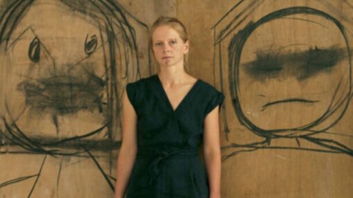 Alt text: A person stands in front of a brown wooden wall with two large, rough black charcoal-style drawings on either side. Each drawing shows an abstract face with exaggerated circular eyes and simple mouth shapes. The person, dressed in dark clothing, is centred between the two expressive, childlike sketches.